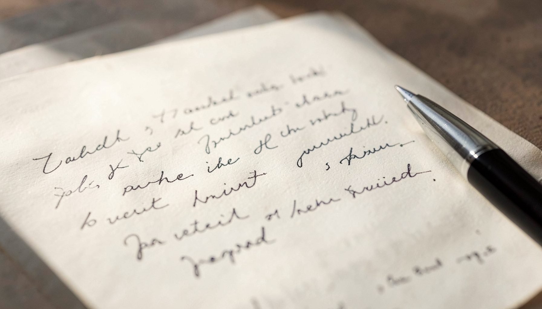 A close-up of a handwritten letter on paper beside a pen, warm natural light, soft cream and muted gray tones